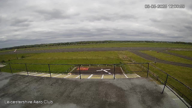 An outdoor view from a webcam showing a cloudy sky over an open grassy area. In the foreground, there is a small parking lot with white lines and an arrow on the ground, and a black railing. To the left, a windsock is mounted on a pole, indicating wind direction. The scene appears to be part of Leicestershie Aero Club, as indicated by a label in the bottom left corner. The timestamp in the top right corner reads "08-05-2025 12:00:58".