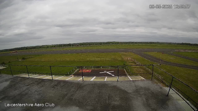 A wide view from a webcam at Leicestershire Aero Club shows an overcast sky with grey clouds. In the foreground, there is a metal railing and a small area with what appears to be marked parking spots or designated zones. Beyond, there is a grassy open field with a track or runway area in the midground. The horizon stretches out with distant greenery and low hills. The timestamp on the top right shows "08-05-2025 12:59:57."