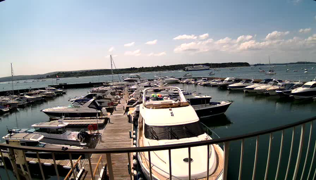 A marina with a wooden pier extending into the water, lined with numerous boats and yachts on both sides. In the foreground, a large white yacht is docked, and behind it, other boats are moored. The water is calm, reflecting the boats. The sky is partly cloudy with a few white clouds, and there is a distant shoreline with some greenery visible on the horizon.