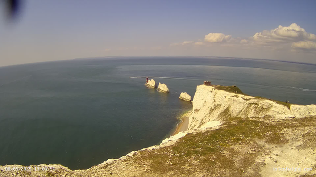 A panoramic view of white chalk cliffs along the coastline, with three prominent sea stacks rising from the water. A small building and a viewing platform are on top of the cliffs. The ocean is calm with subtle waves, and a boat is seen leaving a white trail in the distance. The sky is mostly clear with some fluffy clouds. The timestamp at the bottom left indicates the date and time.