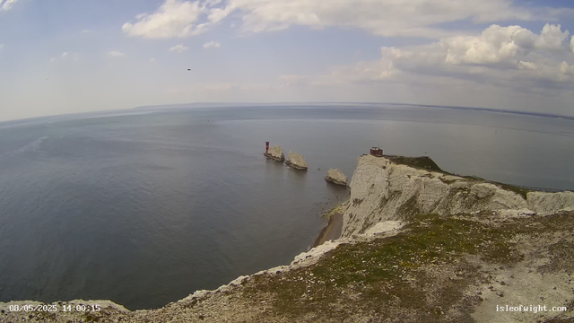 A panoramic view of White Cliffs of Dover, showing tall white chalk cliffs overlooking the English Channel. Several large rock formations are present in the water near the cliffs, including a small lighthouse on one of the rocks. There is a building or structure on top of the cliffs, and a railing along the edge. The sky is partly cloudy with patches of blue. The image has a timestamp at the bottom left corner, indicating it was taken on August 5, 2025, at 14:00:15.