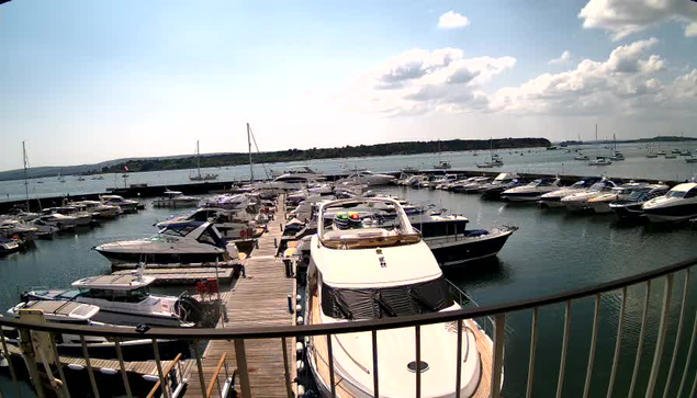 A view of a marina with multiple boats docked along wooden piers. The water is calm, and the sky is partly cloudy with some sunshine. In the background, there is a distant shoreline with greenery. The photo appears to be taken from a balcony or platform with a metal railing in the foreground.