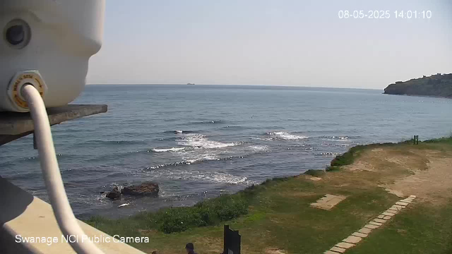 A view of the ocean with gentle waves reaching the shore. In the foreground, part of a white webcam device is visible on the left side. The shoreline has a grassy area and a stone pathway leading towards the water. The sky is clear, and there is a headland with trees on the right side in the distance. The timestamp in the top right corner indicates the date and time as August 5, 2025, at 14:01:10. The bottom left corner shows the label "Swanage NCI Public Camera."
