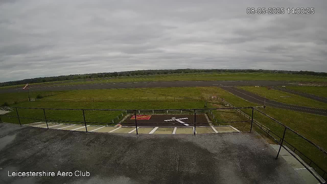 A view from a webcam overlooking a flat, open area with grassy fields and a small runway under a cloudy sky. In the foreground, there is a concrete platform with a metal railing, and a sign indicating the Leicesterhire Aero Club. The timestamp shows the date as August 5, 2025, and the time as 14:00:25.
