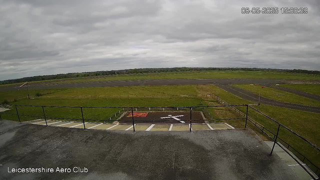 A view from a webcam at Leicestershire Aero Club showing an overcast sky with grey clouds. The image captures a grassy airfield with a runway in the distance, some fencing, and a parking area with marked spaces in the foreground. The timestamp in the top right corner indicates it was taken on August 5th, 2025, at 3:00 PM.