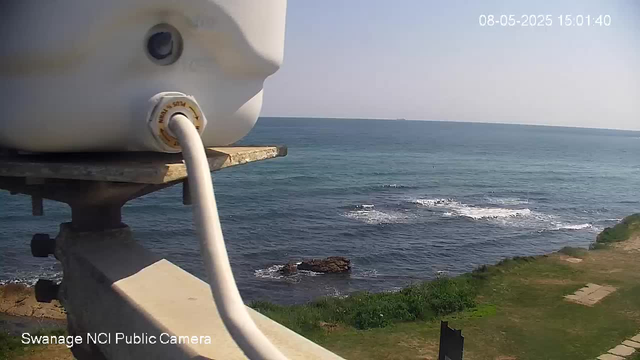 A webcam captures a view of the ocean with gentle waves rolling toward the rocky shoreline and grassy area. The sky is clear and bright, indicating a sunny day. The timestamp in the top right corner shows the date and time as August 5, 2025, at 15:01:40. The camera is mounted on a structure or pole close to the water, with some equipment visible in the foreground.