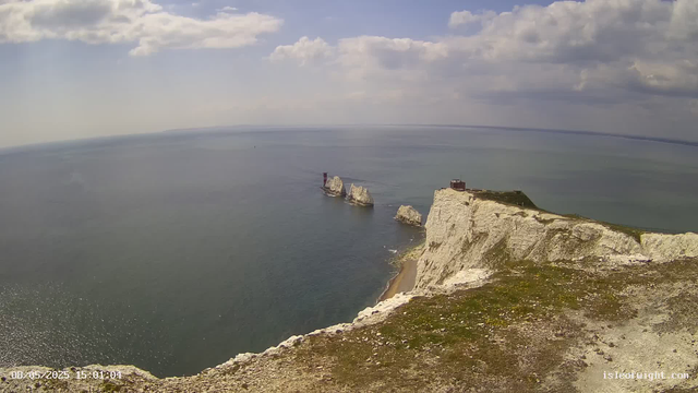 A view of white chalk cliffs meeting the sea, with a lighthouse on a small island or rock formation near the cliffs. The sky is partly cloudy with some sunlight shining through. The water appears calm, and there is a timestamp in the bottom left corner.