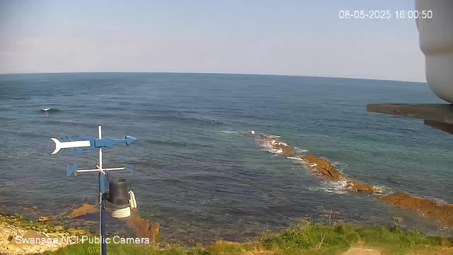 A view of the ocean shoreline with rocky areas, waves gently crashing against the rocks, and some green vegetation at the bottom. There is an anemometer and weather instruments in the foreground, and the sky is clear with a slight haze. The timestamp shows the date and time as August 5, 2025, at 4:00:50 PM.
