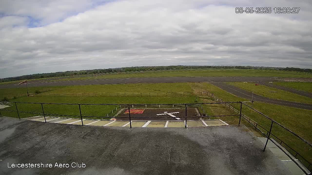 A view from a webcam showing an open runway surrounded by grassy fields under a cloudy sky. There are parking spaces and a small red object in the foreground, with a fence along the edge of the observation area. The timestamp on the image reads "08-05-2025 16:00:27" and the location is labeled as "Leicestershire Aero Club."
