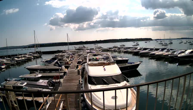 A view of a marina with numerous boats docked along wooden piers extending into the water. The boats vary in size, with some large yachts and smaller vessels. The sky above is partly cloudy, with sunlight breaking through the clouds, reflecting on the calm marina waters. A curved railing is visible in the foreground, indicating the photo was taken from a vantage point or walkway overlooking the marina.