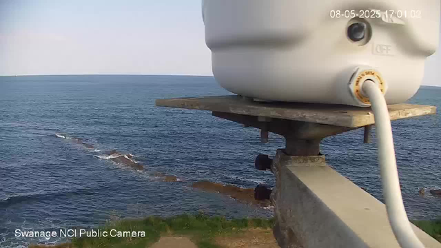 A marine weather station is mounted on a concrete structure overlooking the ocean. The station has a white, rounded device with a small black camera or sensor on its side. A white cable is connected to the device, and the background features a vast view of the ocean with gentle waves under a clear sky. The timestamp on the image reads "08-05-2025 17:01:02," and the bottom left corner indicates it is captured by "Swanage NCI Public Camera."