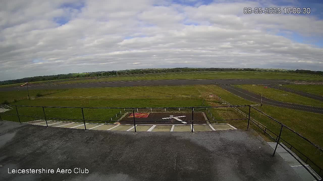 A wide outdoor view from a webcam at Leicester Airport Club, showing a grassy area, a runway in the distance, a small tarmac section with marked parking spaces, and a cloudy sky overhead. The timestamp in the upper right corner reads "08-05-2025 17:00:30."