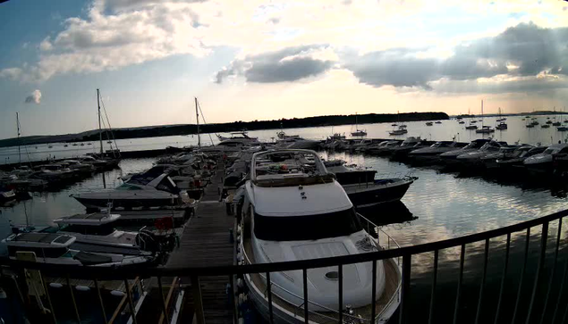A view of a marina with multiple sailboats and motorboats docked along wooden piers. The sky is partly cloudy with the sun setting or rising, casting a soft light on the scene. A metal railing is visible in the foreground.