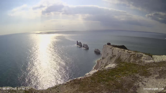 A view from a webcam showing white chalk cliffs overlooking the ocean. The sunlight reflects off the water, creating a shimmering path across the sea. In the distance, there are sea stacks and a lighthouse on one of the stacks. The sky is partly cloudy with rays of sunlight breaking through, and a small boat is visible far out on the water. The timestamp indicates the date and time as August 5, 2025, at 5:00:57 PM.