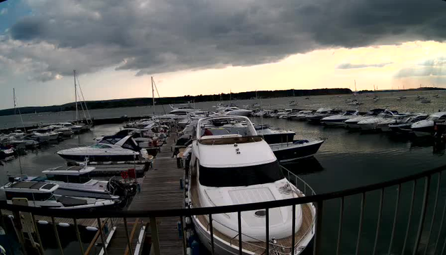 A marina filled with numerous boats docked along wooden piers under a dark, cloudy sky with streaks of sunlight breaking through. The boats vary in size and type, and the water appears calm. The scene is captured from a vantage point behind a black railing in the foreground.
