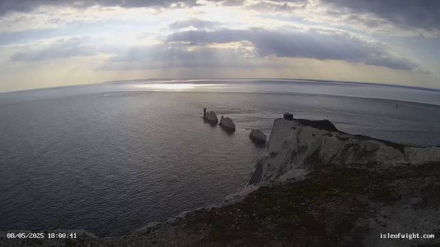 The image shows a view of the ocean from a high vantage point on a cliff. There are several large white chalk rocks protruding from the water near the coast. The sky is partly cloudy with rays of sunlight breaking through the clouds and reflecting off the water. A lighthouse is visible on one of the rocks, and the landscape includes rugged cliffs with patches of grass. The scene appears to be during late afternoon or early evening. The timestamp on the image reads "08/05/2025 18:00:41," and the website is indicated as isleofwight.com.