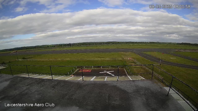 A view of an open grassy airfield under a partly cloudy sky. The image is from a webcam at Leicestershire Aero Club, with a timestamp showing "08-05-2025 18:00:58". In the foreground, there is a parking area with a few marked parking spots and a small helipad with a white arrow pointing to the right. The airstrip extends into the distance, with a runway and taxiways visible. There are some low fences and small structures along the tarmac, and the landscape is mostly flat with a few trees on the horizon.