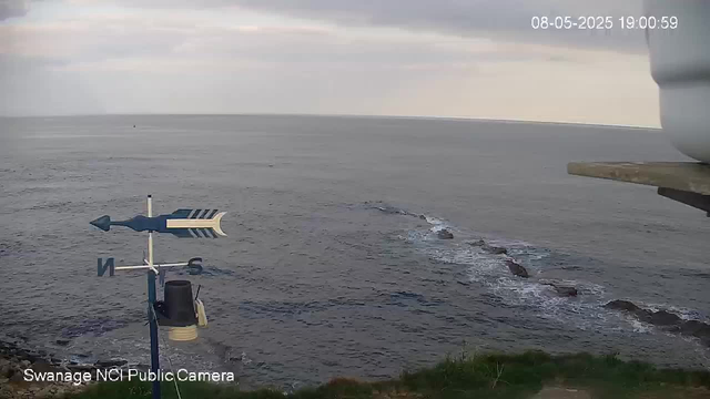 A view of the ocean from a webcam, with a cloudy sky overhead. In the foreground, there is a weather vane with directional markers. Some rocks are visible in the water near the shoreline, which is grassy. The camera timestamp shows the date as August 5, 2025, and the time as 19:00:59. The bottom left corner indicates the camera is a "Swanage NCI Public Camera."