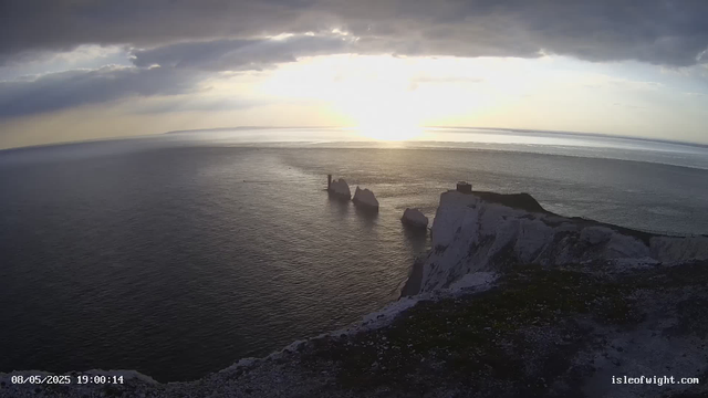A scenic view of white chalk cliffs along the coastline at sunset, with the ocean stretching out below. The sky is partly cloudy, with the sun low on the horizon casting a warm glow over the scene. There are a few large rocks in the water near the base of the cliffs. A small lighthouse or navigation tower is visible on one of the larger rock formations in the sea. The photo is timestamped "08/05/2025 19:00:14" and includes a web address "isleofwight.com" in the bottom right corner.