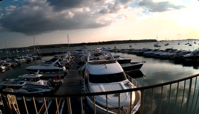 A boat marina with numerous yachts and boats docked along piers. The sky is partly cloudy with the sun setting, casting a warm glow over the water. A curved railing is visible in the foreground.