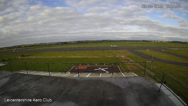A view of an airfield taken from a webcam. In the foreground, there is a rooftop with a railing, and a small parking spot marked with white lines. Beyond that, a grassy area surrounds a runway, with a windsock on the left side. An airplane is on the runway in the distance. The sky is partly cloudy with patches of blue, and the landscape stretches out with fields and trees in the background. The image is timestamped "08-05-2025 19:00:50" and labeled "Leicestershire Aero Club" at the bottom left.