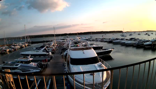 A marina filled with numerous boats docked along wooden piers during sunset, with calm water reflecting the sky and boats, and a distant shoreline visible in the background.