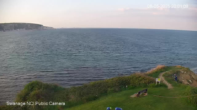 A view of a seaside landscape with a large body of water and distant white cliffs on the horizon. In the foreground, there is a grassy area with a bench and a narrow dirt path leading to a small, rocky outcrop. A person wearing a dark jacket and a helmet is standing on the rocks near a railing. The scene is captured in the evening, with soft light and a clear sky. The timestamp shows the date and time as August 6, 2025, at 8:01 PM.