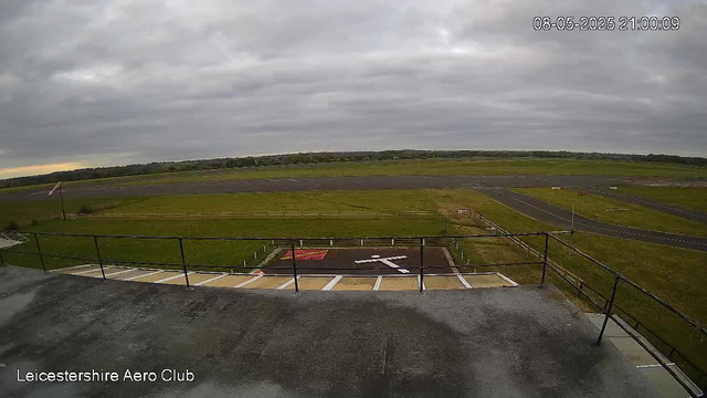 An outdoor view from a webcam showing an overcast sky with grey clouds. In the foreground, there is a concrete platform with a metal railing, and a helipad marked with a white helicopter symbol. Below the platform are designated parking spaces with white lines. In the background, there is a grassy field with a small flag and a distant runway or taxiway. The image is timestamped "08-05-2025 21:00:09" and labeled "Leicestershire Aero Club" in the bottom left corner.
