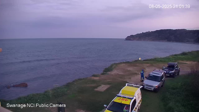 A seaside scene with a grassy shoreline in the foreground and three parked vehicles: a yellow and white ambulance, a silver SUV, and a dark-colored SUV. A person in a blue outfit and helmet is standing near the silver SUV, possibly a worker or emergency personnel. The ocean extends into the distance with gentle waves, and a forested headland juts out into the water on the right side of the image. The sky is mostly cloudy with some sunlight breaking through, creating a calm, overcast atmosphere. The date and time displayed in the upper right corner are August 5, 2025, at 9:01 PM. The bottom left corner shows the label "Swanage NCI Public Camera."