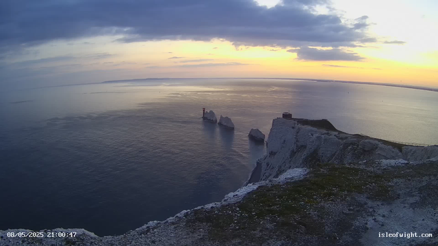 A view of white chalk cliffs along the coastline at sunset, with the sea stretching out to the horizon. There are a few large rocks in the water near the base of the cliffs, and a small building or lighthouse sits atop the highest point of the cliffs. The sky is partly cloudy, with the sun setting behind the clouds, casting a soft glow over the scene. In the bottom left corner, there is a timestamp that reads "08/05/2025 21:00:47," and in the bottom right corner, a website URL "isleofwight.com" is visible.