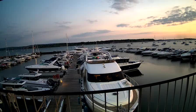 A marina at sunset with many boats docked along wooden piers. The sky is partly cloudy with a soft glow from the setting sun, casting a calm and peaceful atmosphere over the water. The boats vary in size and style, with some larger yachts in the foreground and smaller sailboats and motorboats further out.