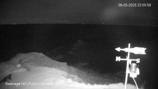 A black-and-white image from a webcam showing a dark, nighttime scene. In the foreground, there is a weather vane with directional markers (N, S, E, W) and a small container attached. The landscape appears to be a sandy or rocky area, with a low hill or dunes to the left. The background is mostly dark with a faint horizon line, and a few small bright spots in the sky, possibly stars or distant lights. The timestamp in the upper right corner indicates the date and time as August 5, 2025, at 10:00:59 PM. The bottom left corner has text stating "Swanage NCI Public Camera."