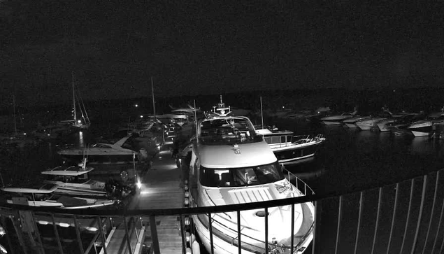 A nighttime view of a marina filled with various boats and yachts docked along the piers. The scene is captured from a balcony with a metal railing at the bottom of the image. The boats are stationary, and the water appears calm. The background shows a dark sky, indicating it is nighttime.