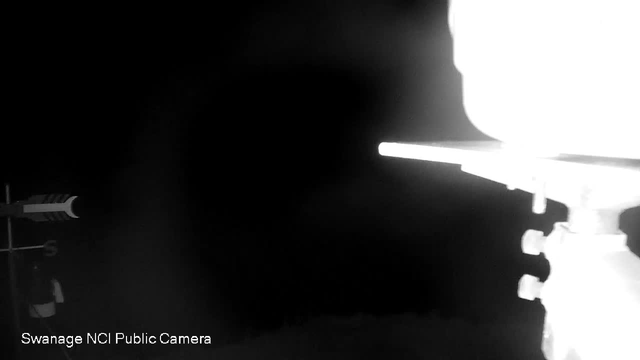 A black and white image from a webcam showing a dark, almost empty room with a bright light source on the right side, illuminating part of the wall and ceiling. In the bottom left corner, there is text that reads "Swanage NCI Public Camera."