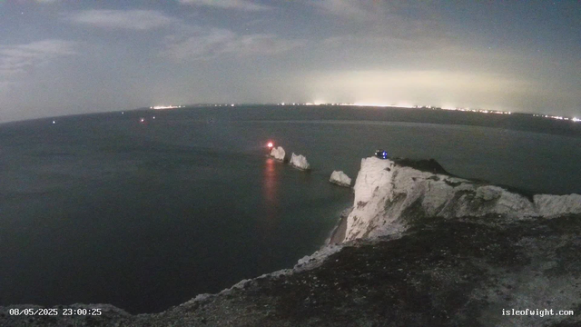 Nighttime view of a coastal cliff with white chalk formations. The sea below is dark with gentle waves, and several navigation lights are visible on rocks in the water. In the distance, there are faint city lights along the horizon. The sky is mostly cloudy with some light glow from the city or possibly atmospheric reflection. The timestamp indicates the photo was taken on August 5, 2025, at 11:00 PM.