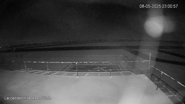 A black-and-white image from a webcam shows an outdoor area at night, with empty parking spaces arranged in front of what appears to be a runway or open space. There is a fence or railing in the foreground, and the sky is dark with some faint stars visible. The timestamp in the top right corner indicates the date as August 5, 2025, at 11:00 PM and 57 seconds. The bottom left corner has text that reads "Leicestershire Aero Club." There is a large bright spot or reflection on the right side of the image.