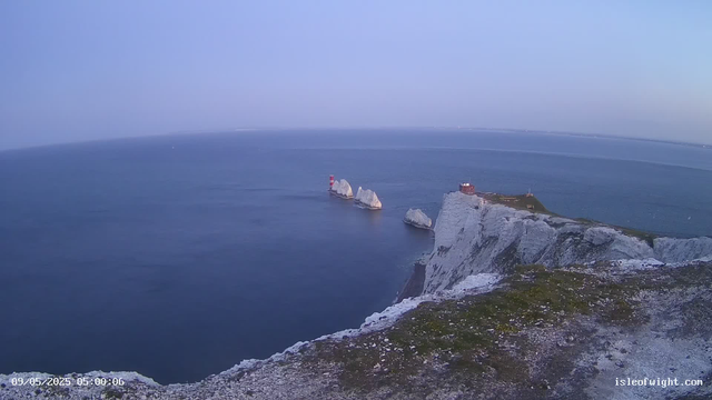 A view from atop the white chalk cliffs of Dover showing a calm blue sea stretching to the horizon. In the water, there are several large white rock formations, with one that has a small red and white lighthouse on top. On the right side of the image, a building and some structures are situated on the cliff's edge. The sky is mostly clear, with a faint haze in the distance. The timestamp at the bottom left reads "09/05/2025 05:00:06" and the bottom right displays "isleofwight.com."