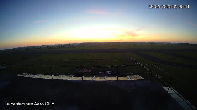 A view from a webcam showing the sky at sunrise with soft orange and pink hues blending into light blue. The landscape features open grassy fields with some fencing and a small runway or pathway in the foreground. The scene is calm and expansive, with the horizon stretching far into the distance. The image is timestamped "09-05-2025 05:00:44" in the top right corner and labeled "Leicestershire Aero Club" in the bottom left corner.