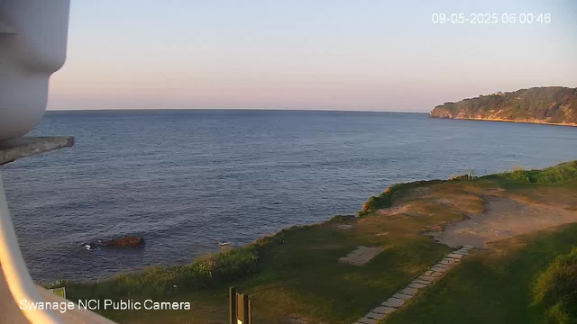 A scenic view of a coastal landscape at sunrise or sunset. The image features calm ocean waters extending to the horizon, with a distant land mass covered in trees on the right. In the foreground, there is a grassy area with a stone pathway leading toward the water, and a large rock near the shoreline. The scene appears peaceful with clear skies. The timestamp in the top right corner indicates it was captured on September 5, 2025, at 6:00 AM.