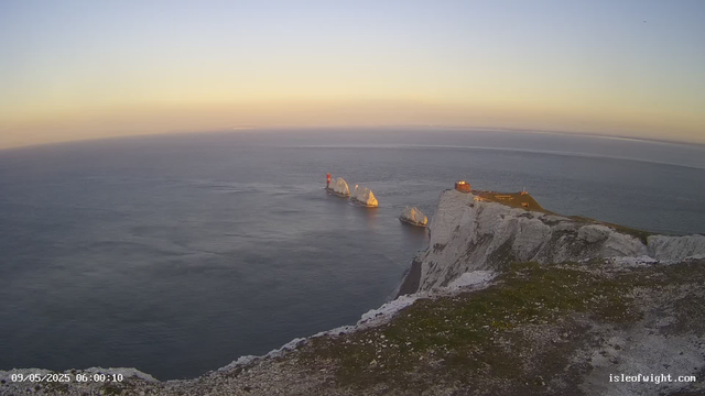 Aerial view of white chalk cliffs with grassy tops along the coastline at sunrise. The sea below is calm with gentle waves, and a lighthouse stands on one of the rocky outcrops in the water. The timestamp shows "09/05/2025 06:00:10" and the bottom right corner displays "isleofwight.com".