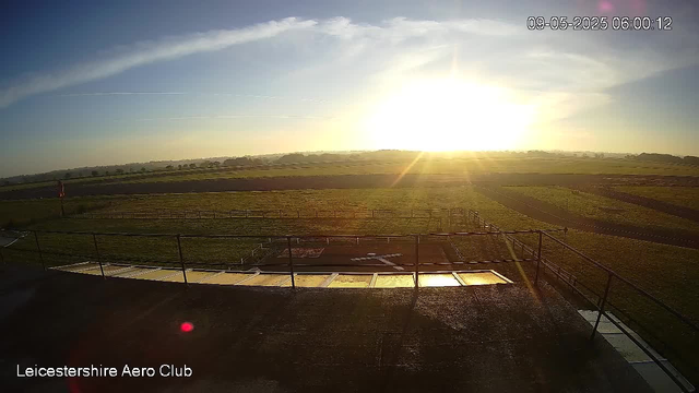 A view of an open grassy field with a track, taken from a webcam at the Leicesterhire Aero Club. The sun is low on the horizon, creating a bright glare and long shadows across the landscape. The sky is mostly clear with a few wispy clouds and a couple of contrails. The image is timestamped "09-05-2025 06:00:12". Railings and a small platform are visible in the foreground.