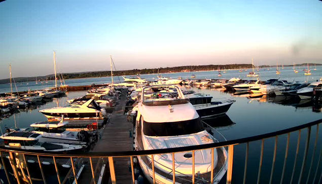 A marina with multiple boats docked along wooden piers. The calm water reflects the boats and the sky, which is clear with a soft gradient from light blue to warm hues near the horizon. In the background, there is a stretch of land or island with trees. The scene is bathed in warm, late-afternoon or early-evening light.