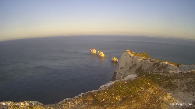 A view of white chalk cliffs extending into the ocean, with a small building on top of the cliff. Several large rock formations are visible in the water near the cliffs, and the scene is bathed in golden sunlight, suggesting early morning or late afternoon. The sky is clear and the water is calm. The timestamp on the image reads "09/05/2025 07:00:29," and there is a website URL "isleofwight.com" in the bottom right corner.