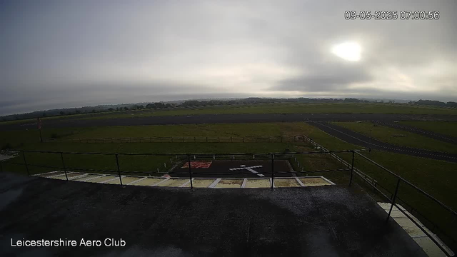 A view from a webcam at Leicestershire Aero Club showing an overcast sky with the sun partially obscured by clouds. The scene includes an open grassy area with a small helicopter landing pad marked with a white helicopter symbol, surrounded by a black border. There is a railing on the foreground terrace, and the background features a flat landscape with fields, trees, and roads stretching into the distance. The timestamp reads 09-05-2025 07:00:56.