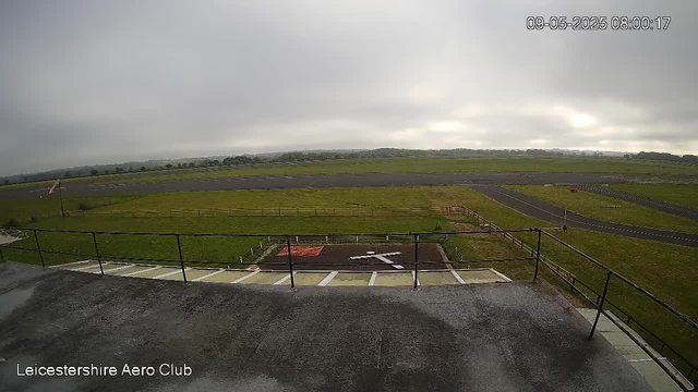 A view of an empty outdoor space at Leicesterhire Aero Club on a cloudy day. The foreground shows a concrete area with some parking spots and a metal railing. Beyond that, there is a grassy field with a small section of fencing and a windsock. The background features a vast open landscape with a runway and distant trees under a gray, overcast sky. The timestamp in the top right corner reads "09-05-2025 08:00:17."