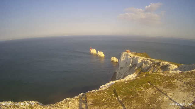 A view from a webcam showing white chalk cliffs overlooking the ocean. Several large rocks and a red and white lighthouse are visible in the water near the cliffs. The sky is mostly clear with a few clouds, and the scene is bathed in warm sunlight. The timestamp in the lower left corner indicates the date and time as 09/05/2023 at 08:00:48.