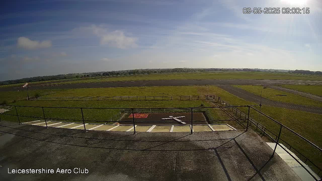 An outdoor view from a webcam showing a bright, partly cloudy sky over a flat, grassy landscape with some paved areas and a small fenced section in the foreground. There's a railing along the edge of the platform where the camera is positioned, and a distant horizon with low-lying trees and fields. The image is labeled "Leicestershire Aero Club" at the bottom left corner, and the timestamp in the top right corner indicates "09-05-2025 09:00:15."
