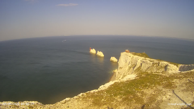 A view of a coastal cliff with white chalk formations extending into the ocean. There is a small building and some structures at the top of the cliff. In the water, three large white rock formations are visible, with a lighthouse on one of them. The scene is captured during daylight with clear skies.