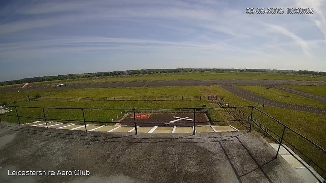 The image shows a wide view of an airfield from a CCTV camera, with a fenced area in the foreground, a grassy open space, taxiways, and runways stretching towards the horizon. The sky is mostly clear with some wispy clouds, and a small flagpole with a flag is visible on the left. The timestamp in the upper right corner reads "09-05-2025 10:00:05," and the bottom left corner indicates it is at the "Leicestershire Aero Club."