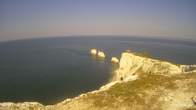 A panoramic view of white chalk cliffs along the coastline under a clear blue sky, with a large cliff in the foreground featuring a small building or lighthouse on top. In the distance, there are several white chalk stacks emerging from the sea, with one topped by a red and white lighthouse. The ocean appears calm beneath the cliffs and stacks, and the horizon stretches far into the distance.